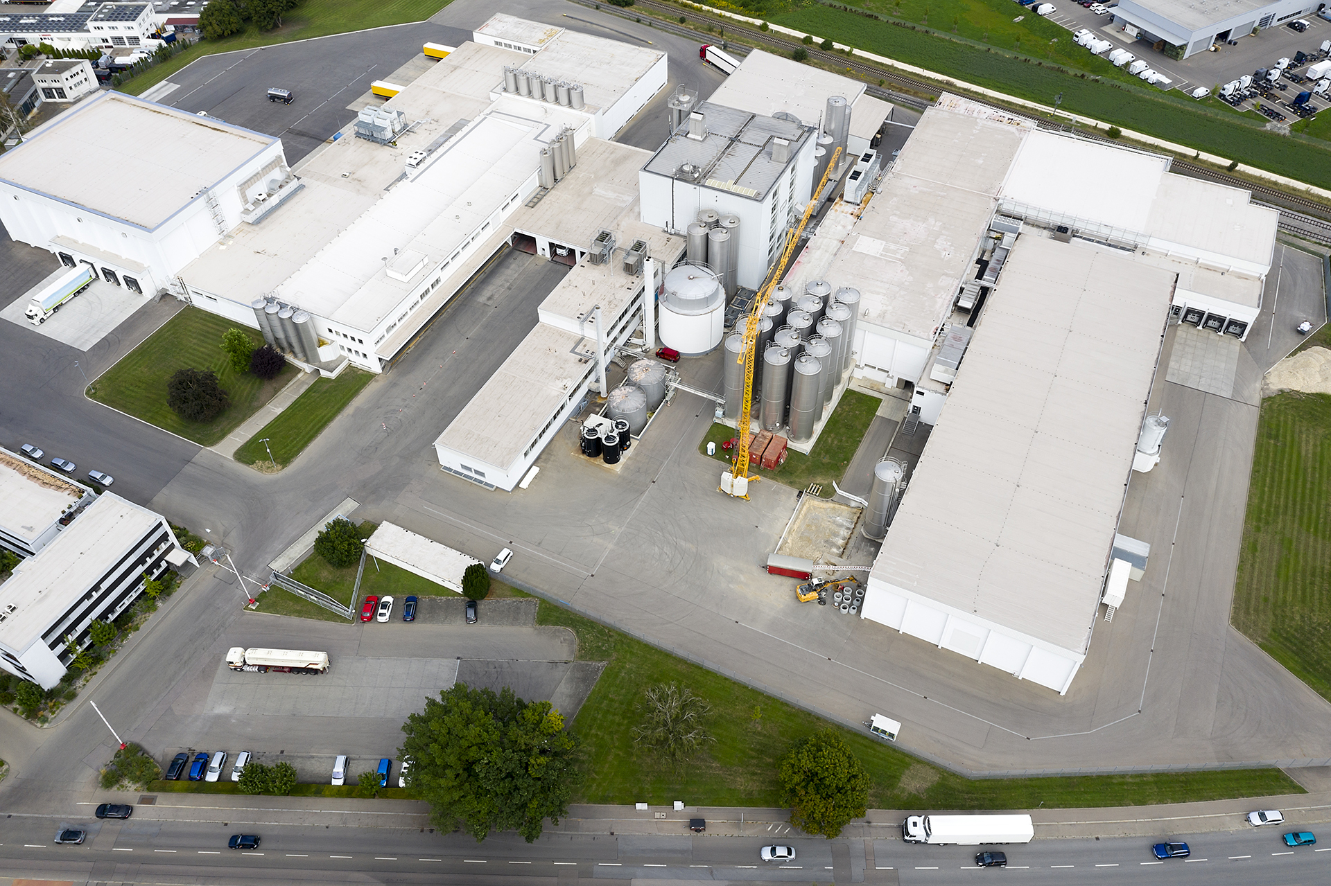 Aerial view of an industrial complex with large white buildings, cylindrical storage tanks, and parked trucks. Surrounding roads and greenery are visible, conveying a sense of scale and organization.