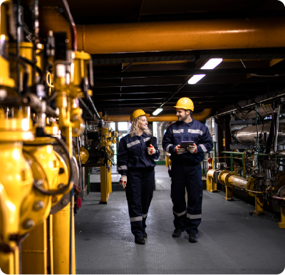 Two factory workers in safety equipment walking by gas pipes in an oil and gas refinery